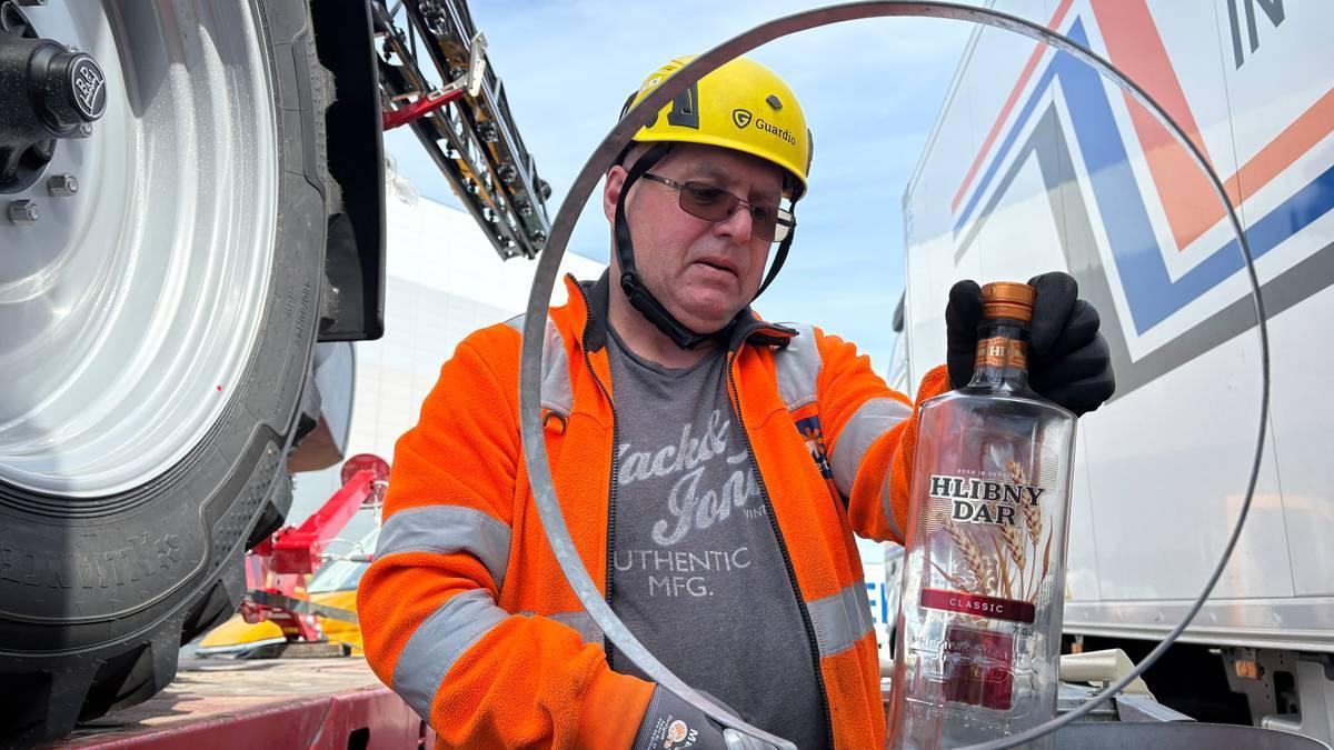 Foto: Anne Ognedal / NRK Un uomo con un casco giallo e una giacca arancione è all’aperto e mostra una bottiglia di superalcolico vuota; accanto a lui c’è un camion e, dall’altro lato, si vede una grande ruota di trattore