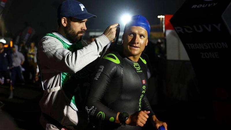 Foto: MELINA PIZANO / GETTY IMAGES NORTH AMERICA,NTB Un uomo in muta viene aiutato da un altro uomo. L’uomo in muta indossa una cuffia da nuoto e ha una bandiera norvegese sul braccio. Dietro di loro ci sono delle persone e un cartello con la scritta «TRANSITION ZONE». (La descrizione dell’immagine è stata realizzata da un servizio di IA)