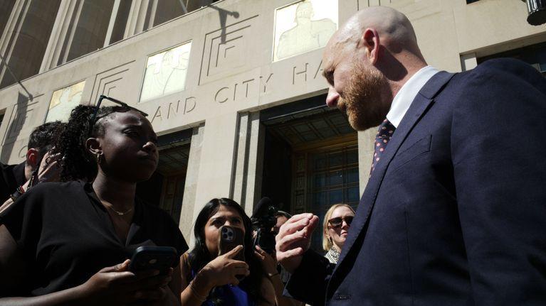None Diverse persone sono in piedi all’esterno, davanti a un edificio; un uomo in abito elegante parla con il gruppo. Sullo sfondo si legge parzialmente «CITY HALL».