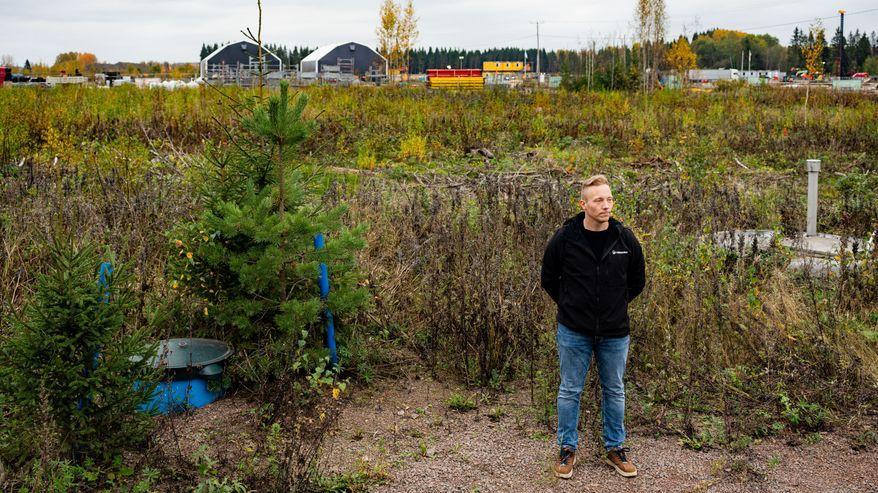 Un uomo è in piedi sul bordo di una strada sterrata grigia, tra la vegetazione; sullo sfondo si vedono edifici industriali e un autobus giallo.