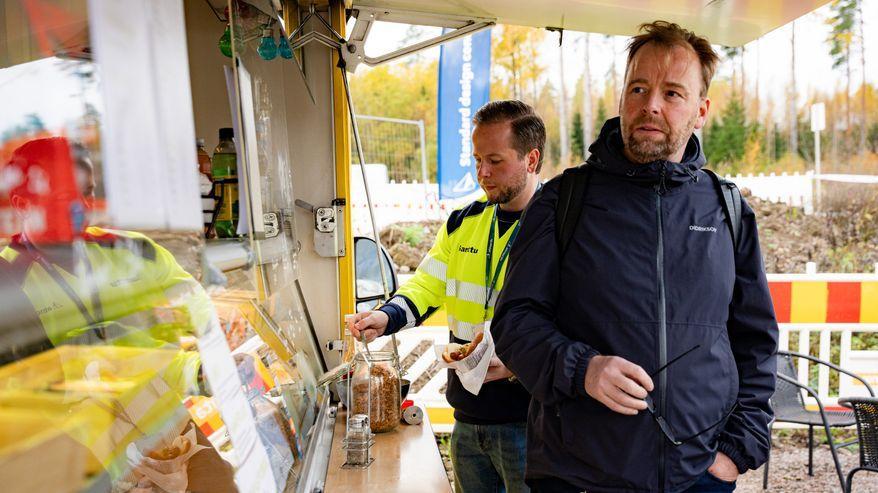 Due uomini sono davanti a un chiosco gastronomico; uno indossa un giubbotto ad alta visibilità e tiene in mano una ciotola di cibo.