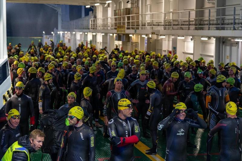 Foto: Amalie Straume / NRK Partecipanti in muta e cuffia da bagno gialla riuniti sul ponte di un traghetto prima del triathlon Norseman. (La descrizione dell’immagine è stata generata da un servizio di IA)