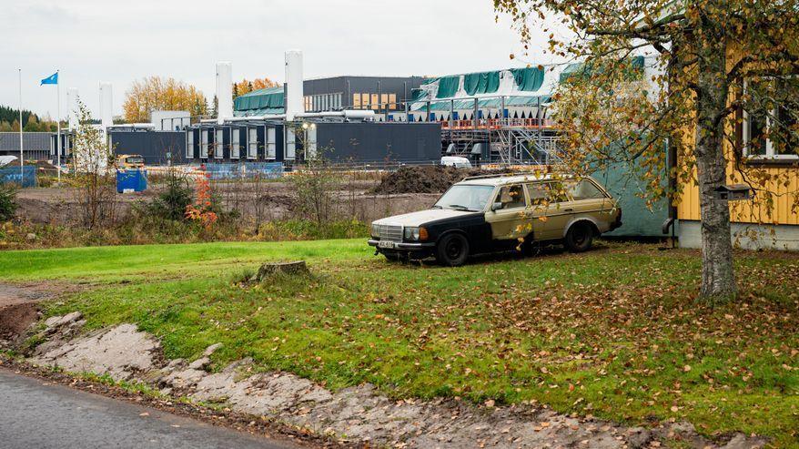 Un’auto piuttosto vecchia è parcheggiata su un prato coperto di foglie, accanto a un edificio giallo; sullo sfondo si vede il cantiere di un data center.