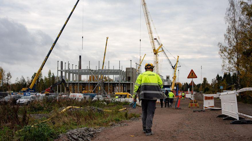 Un cantiere con diverse gru e un edificio incompiuto; in primo piano una persona in abiti da lavoro cammina.
