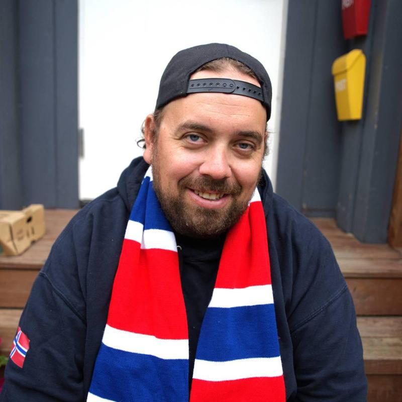 Foto: Mathias Nordhus-Kristoffersen / NRK Un uomo è seduto su una scala davanti a una porta bianca, con felpa con cappuccio, cappellino e sciarpa da tifoso rossa, bianca e blu. Ha la barba e sorride alla fotocamera.